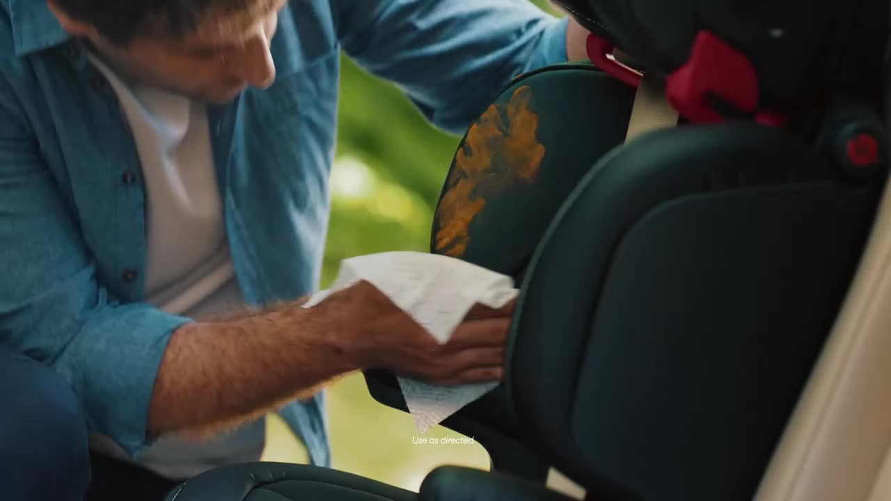 A man wearing a blue shirt is wiping a stained car seat with a white wipe. The seat has a large, orange-brown stain on the headrest.
