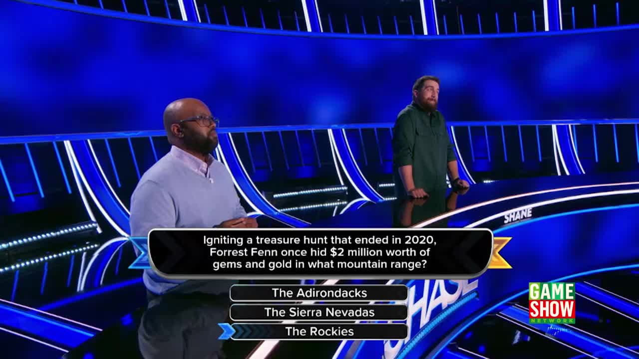 On a Game Show Network set, two men stand on a brightly lit stage. A question about a treasure hunt is displayed, and one contestant looks thoughtful while the other waits.
