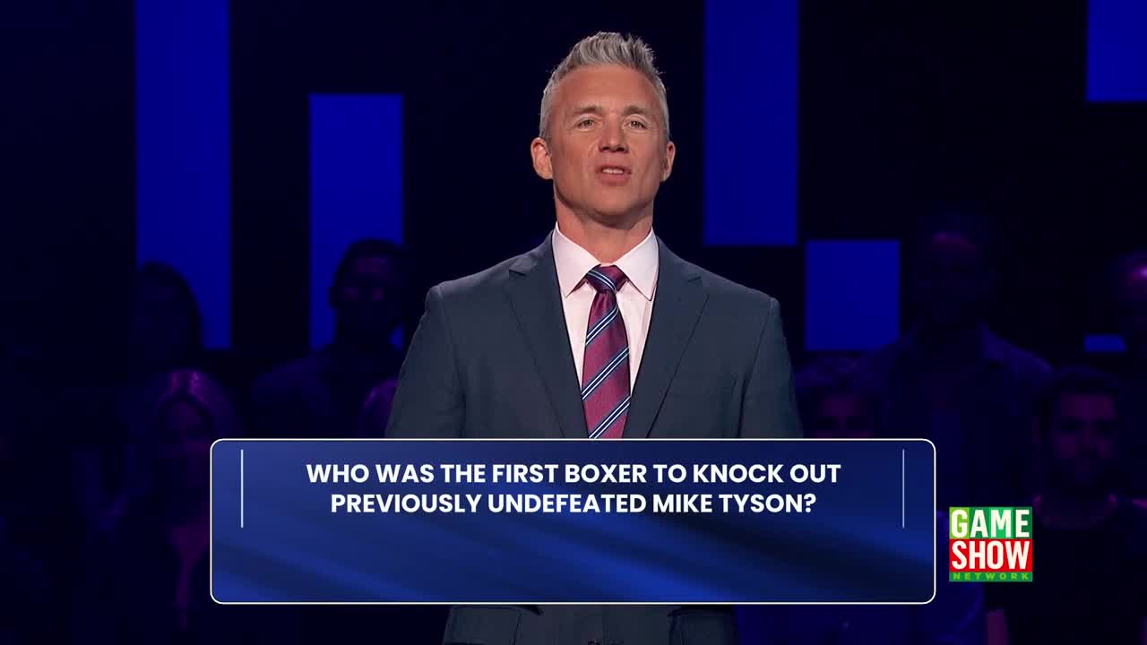 A man in a suit, with silver hair, is speaking to the camera. Below him, a question about Mike Tyson is displayed on a blue screen, with the Game Show Network logo visible on the right.
