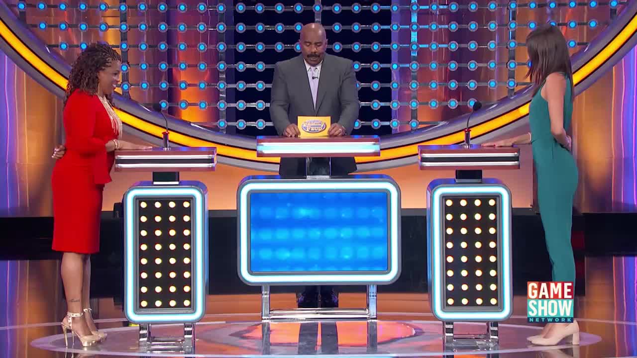 Steve Harvey stands center stage, holding a yellow game card. Two women, one in red and one in teal, face him from their podiums. The Game Show Network logo is visible on the right.