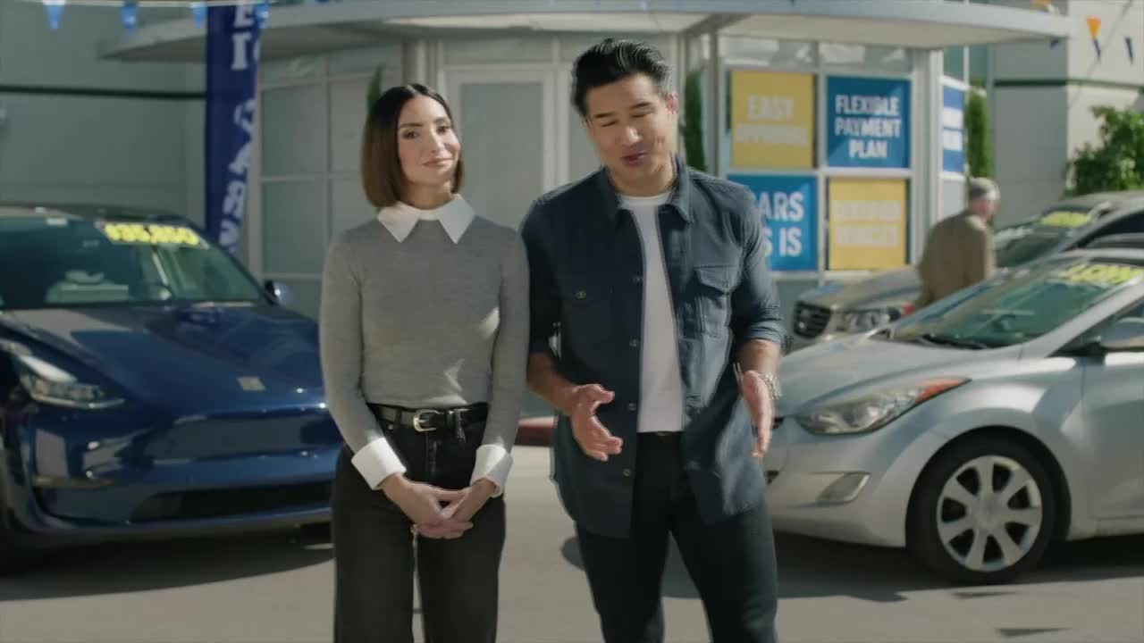 A man and woman stand in front of a row of cars at a dealership. The man gestures with his hands as he speaks, while the woman stands with her hands clasped.