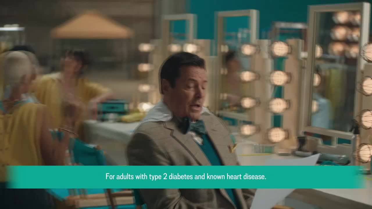 A man in a suit looks down at papers, his mouth slightly open. In the background, people in yellow shirts are moving around a set with bright lights.