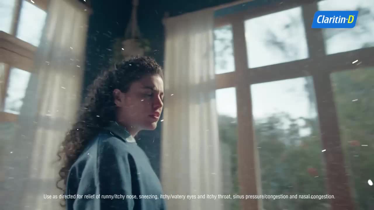 A woman with curly hair turns her head, her face showing discomfort as tiny particles float in the air around her. The light from the window catches these specks, making them visible against the dim interior.