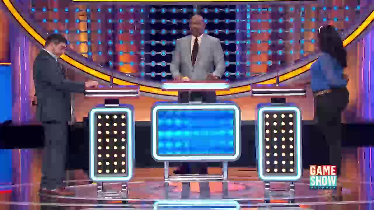 A man in a suit stands at a podium, his hand hovering over a button. To his right, a woman in a blue shirt also stands, facing away. The host, in a light grey suit, stands between them, looking at the blue screen in front of him.