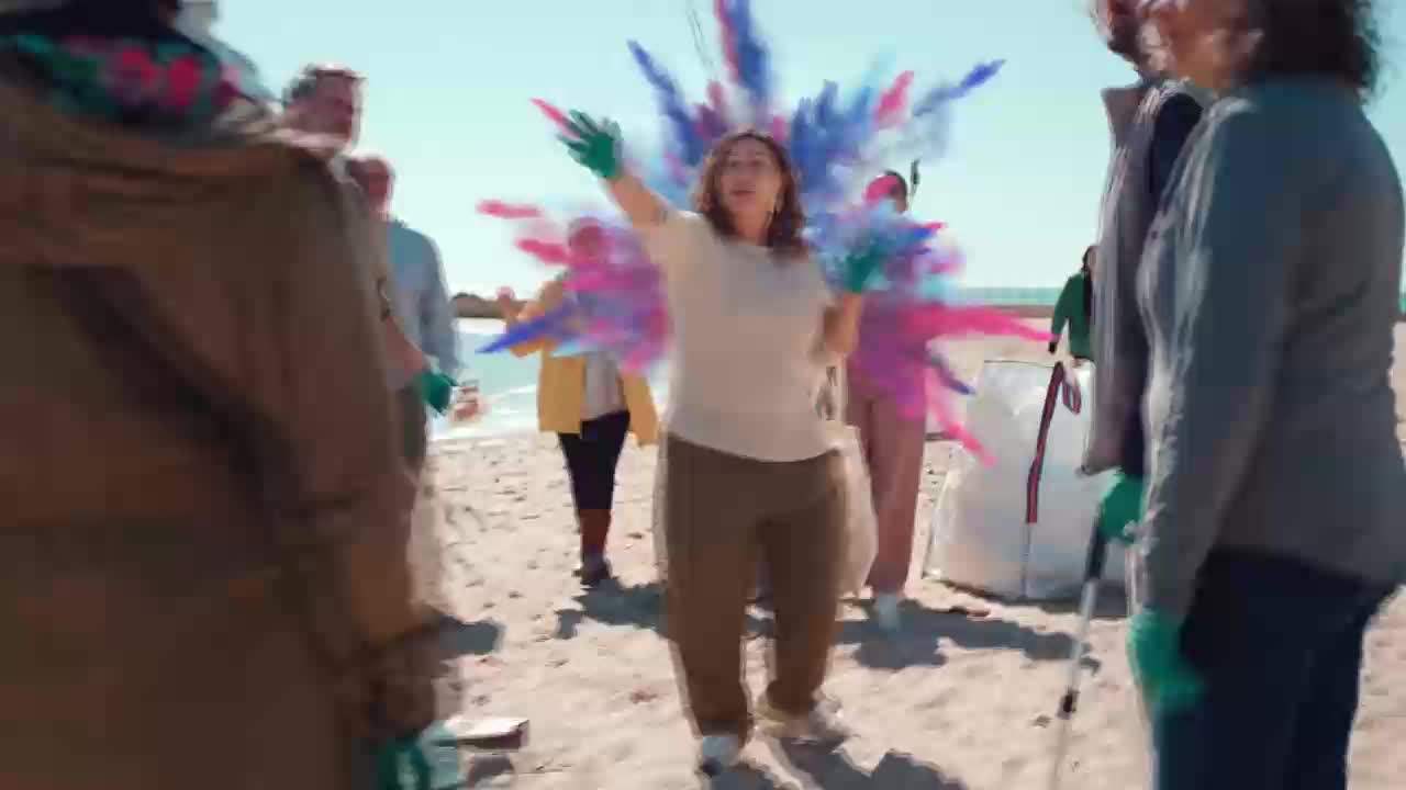 A woman in the center throws a cloud of pink and blue powder into the air. A group of people stand on a beach, some wearing gloves.