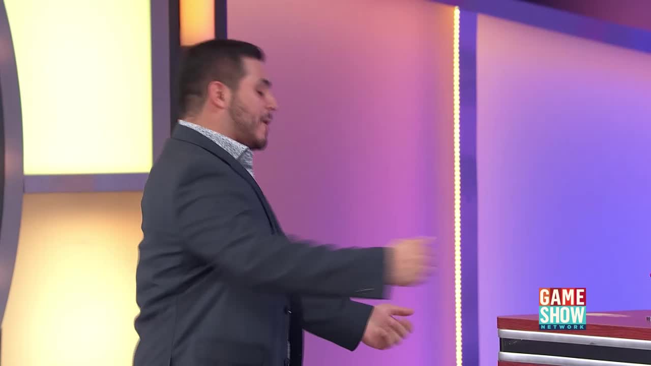 A man in a dark suit gestures with his hands, his arms blurred from movement. He stands on a brightly lit stage, the Game Show Network logo visible on a nearby podium.