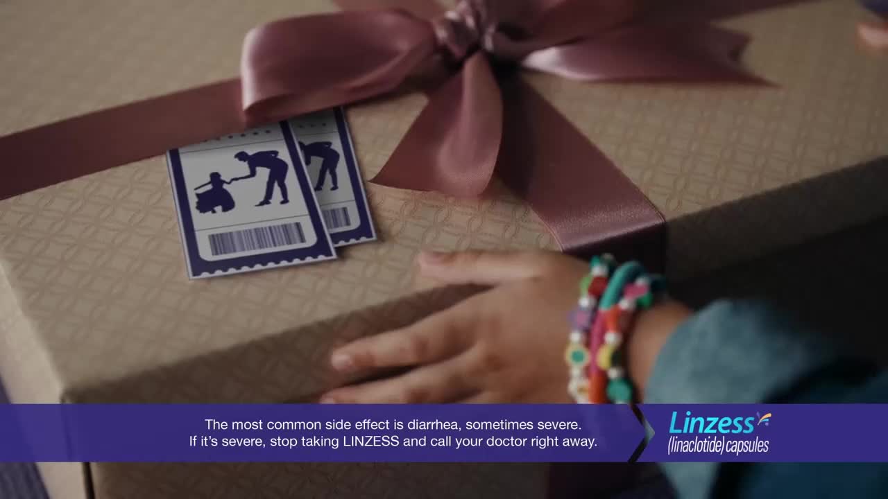 A child's hand, adorned with colorful bracelets, reaches for a gift box tied with a pink ribbon. Two small blue tickets with silhouette figures are tucked under the ribbon.