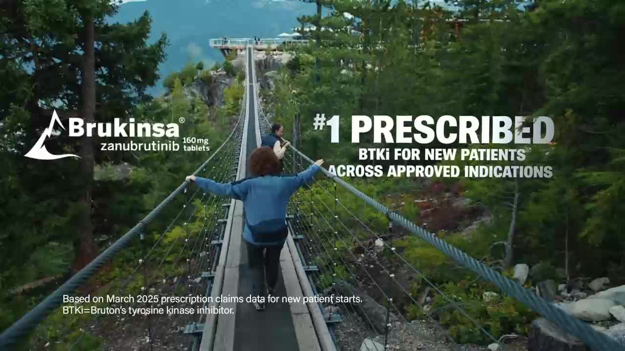 Two people walk across a suspension bridge high above a forested valley. The bridge is made of wood planks and thick steel cables, with a railing to hold onto.