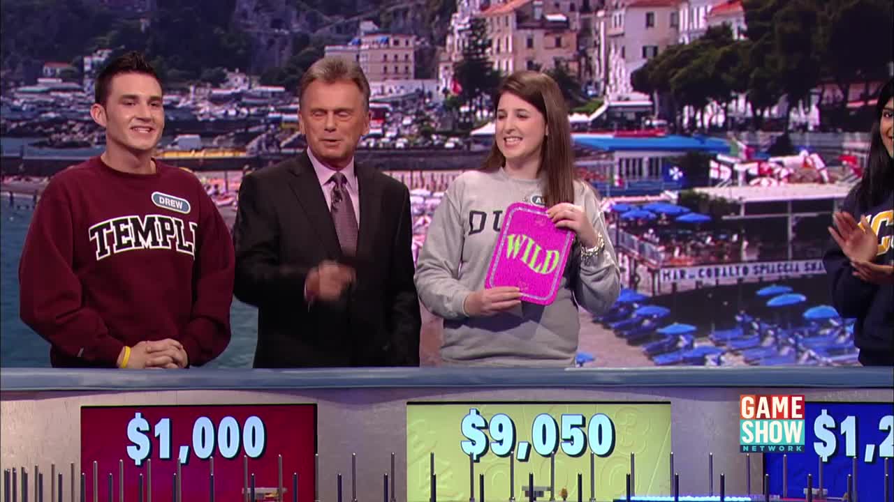 The host gestures as a contestant on the Game Show Network holds up a pink "WILD" card. Another contestant smiles, wearing a maroon sweatshirt with "TEMPLE" across the chest.