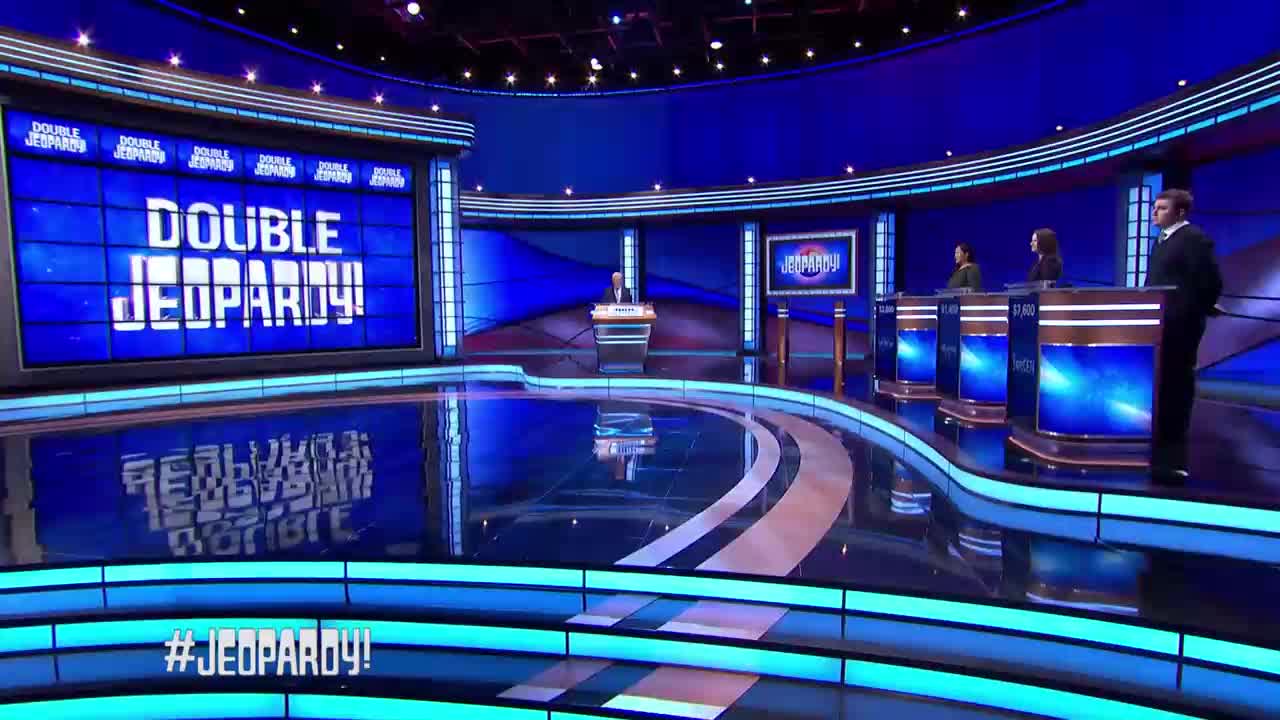 The host stands at his podium as three contestants wait behind their stations. A large screen behind them displays "DOUBLE JEOPARDY!" in bright white letters.