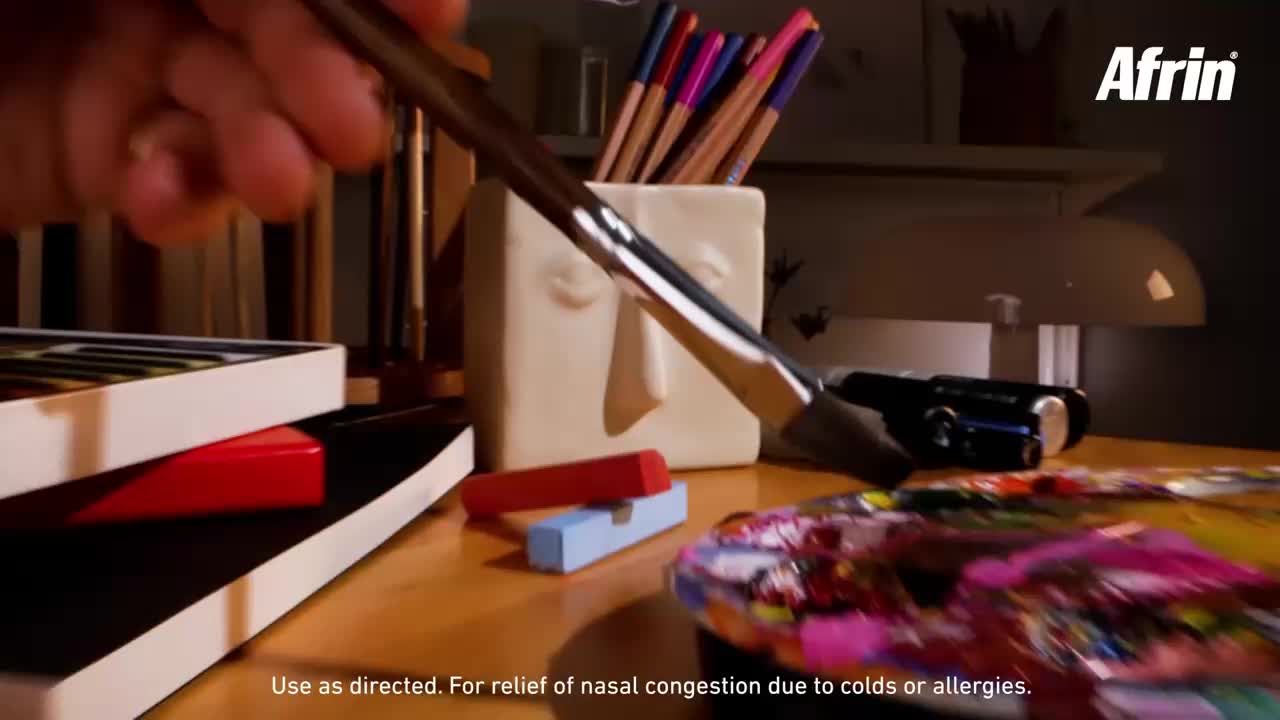 A hand dips a paintbrush into a palette of vibrant colors. Pencils stand upright in a quirky face-shaped holder on the desk.