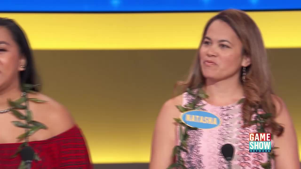Two women stand on a brightly lit stage, both wearing leis. The woman on the right, Natasha, smiles as she looks off-camera. A Game Show Network logo appears on her right.