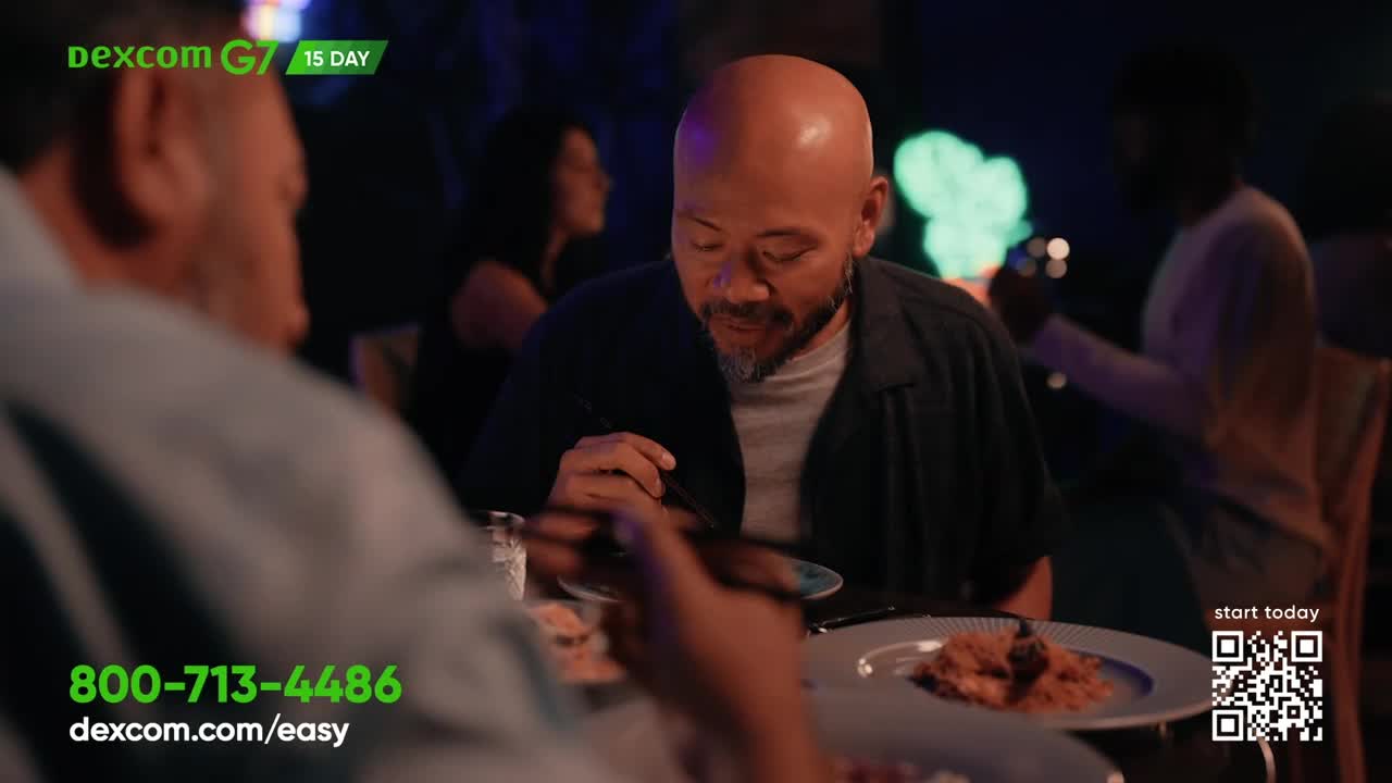 A man with a shaved head eats from a plate with chopsticks, his gaze lowered. In the background, other diners are visible in the dim, atmospheric lighting.