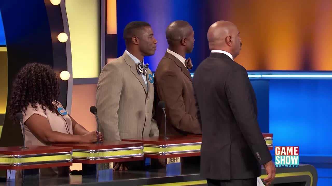 Three men in suits stand at podiums on a brightly lit game show stage. A woman with curly hair sits at a podium to the left, facing away from the camera.
