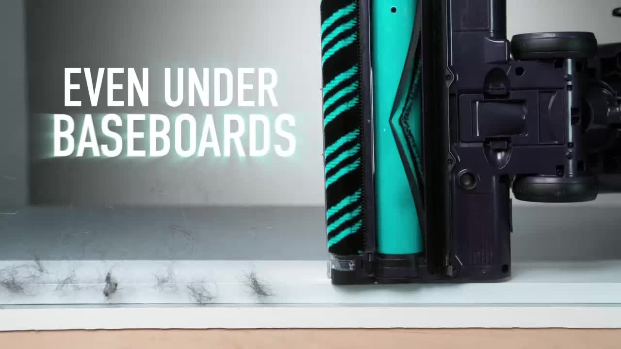 A vacuum cleaner's brush head, with its teal and black bristles, glides along a white surface. Dust bunnies and hair are being pulled into the vacuum from the edge of the baseboard.