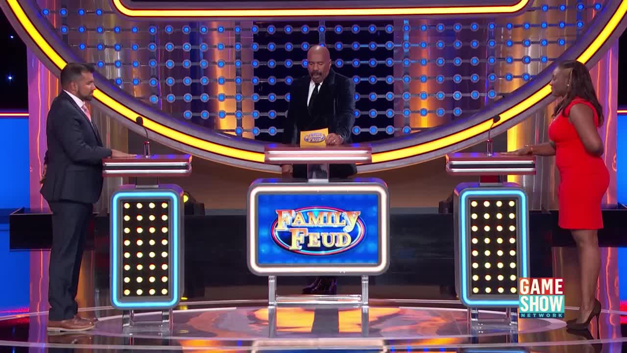 Steve Harvey stands center stage, holding a card, as two contestants face him. The bright lights of the Family Feud set illuminate the scene.