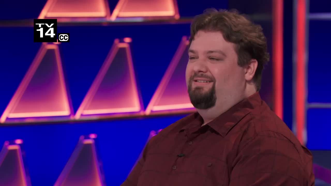 A man in a maroon shirt smiles, his gaze directed off-camera. Behind him, the Game Show Network's signature triangular set glows with purple and blue lights.