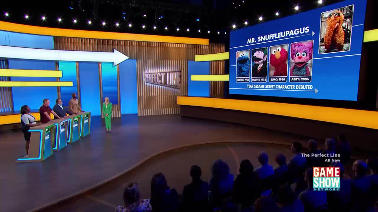 A host in a bright green suit stands facing a line of contestants at their podiums. On a large screen behind them, images of Sesame Street characters, including Mr. Snuffleupagus, are displayed with their debut years.