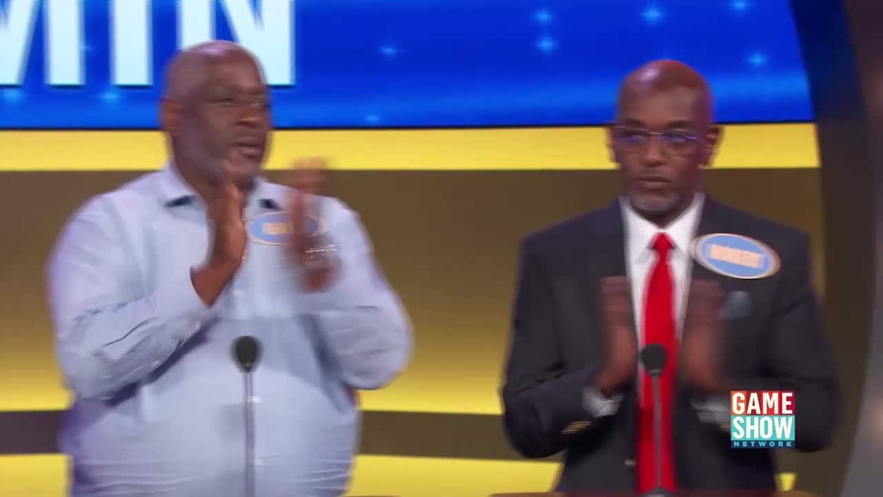 Two men stand at microphones, clapping their hands together. The man on the left wears a light blue button-down shirt, while the man on the right is in a dark suit with a bright red tie. A logo for the Game Show Network is visible in the lower right corner.