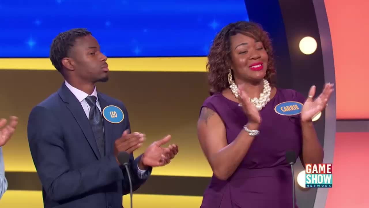 Leo, in his dark suit, claps his hands together with a focused expression. Beside him, Carrie, wearing a vibrant purple dress, smiles and claps, her pearl necklace catching the light.