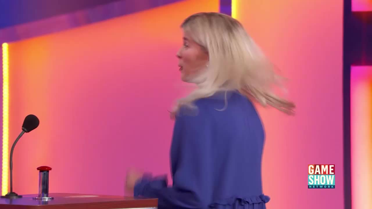 A woman in a blue dress spins, her blonde hair flying out. The bright pink backdrop glows behind her.