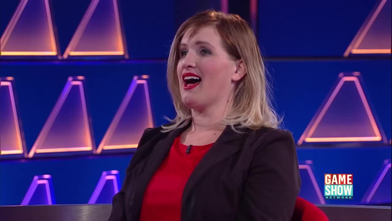 A woman in a red shirt and black blazer is speaking, her mouth open in excitement. Behind her, triangular lights glow on a blue backdrop, a familiar sight from the Game Show Network.