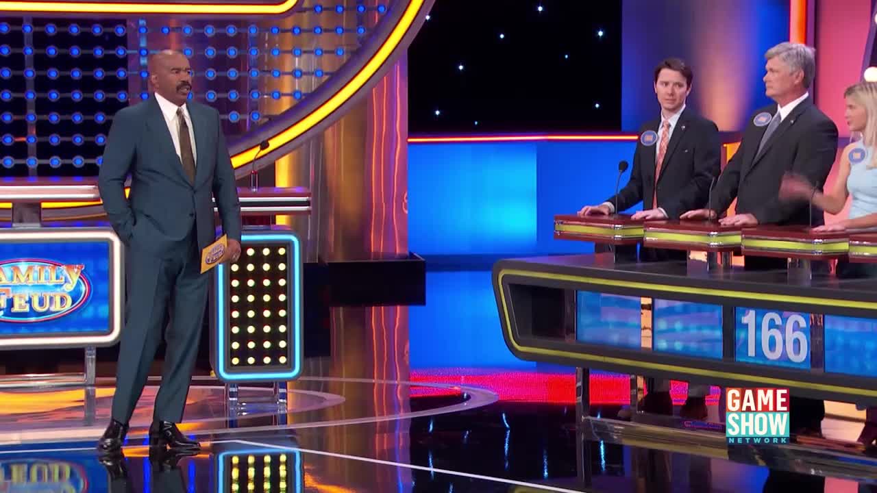 Steve Harvey stands on the left, holding a yellow card. Three contestants in suits stand at their podiums on the right, facing forward.