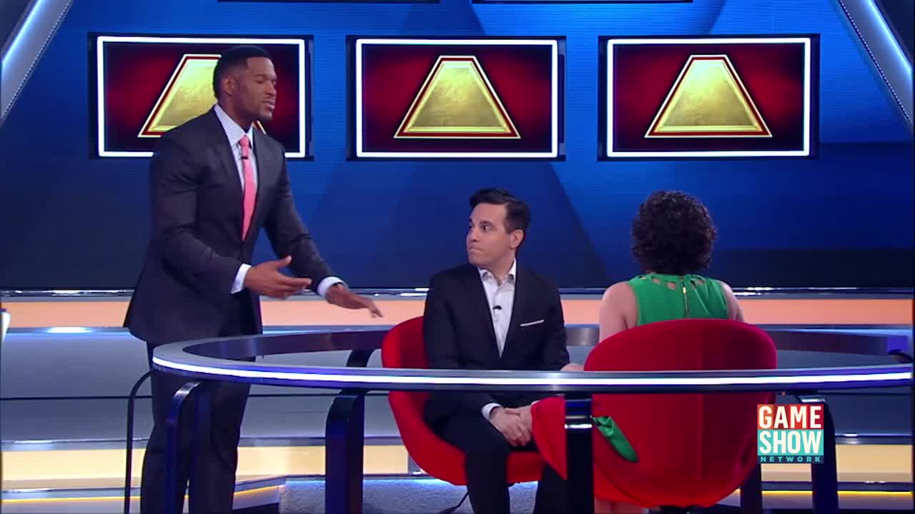 The host, Michael Strahan, gestures toward the contestants as they sit in their red chairs. Three golden triangles illuminate the blue backdrop behind them, a familiar sight on the Game Show Network.