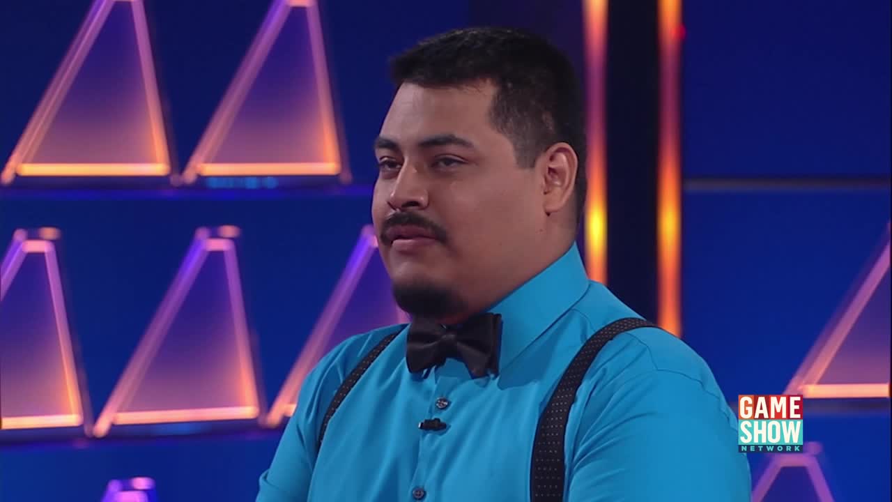 A man in a bright blue shirt and black bowtie looks off to the side. Behind him, illuminated triangles glow in shades of orange and purple.