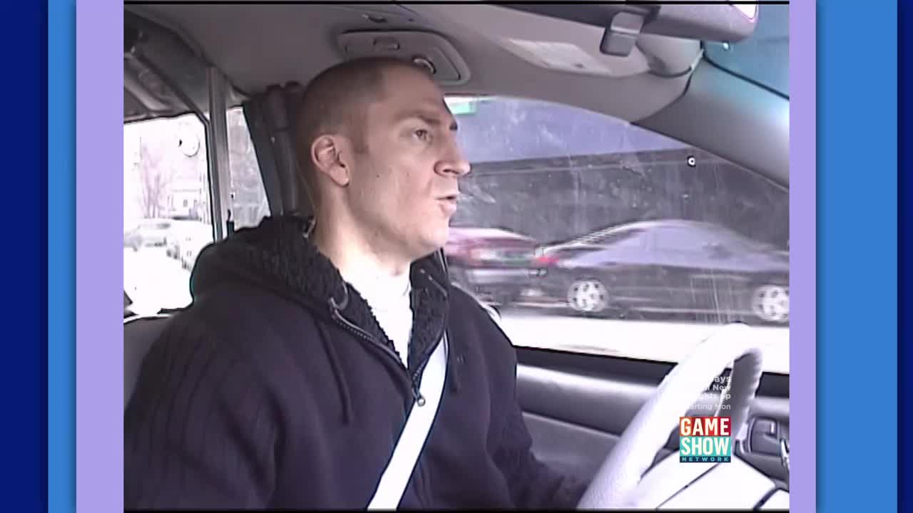 A man grips the steering wheel, his eyes focused ahead as cars pass by outside the window. A "Game Show Network" logo is visible on the dashboard.