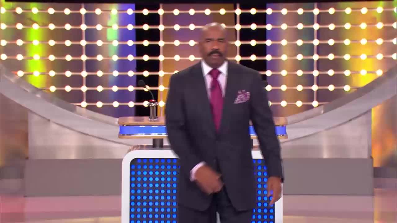 Steve Harvey, host of the popular game show, walks across the stage. The colorful, illuminated backdrop of the Family Feud set shines behind him.