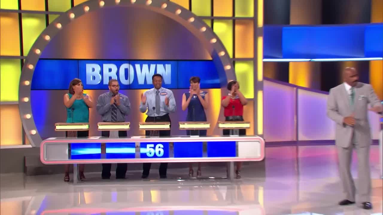 The "Brown" family stands at their podiums, hands to their faces in shock or surprise. Steve Harvey, in a light gray suit, walks across the stage towards the contestants.