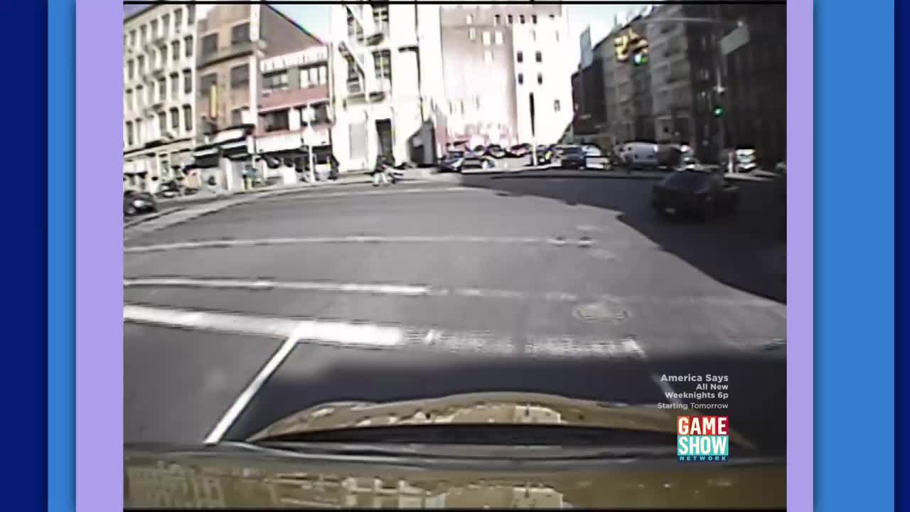 A motorcycle is turning left at a busy intersection in a city. Cars are stopped at the red light, and a dark sedan is driving away from the camera.