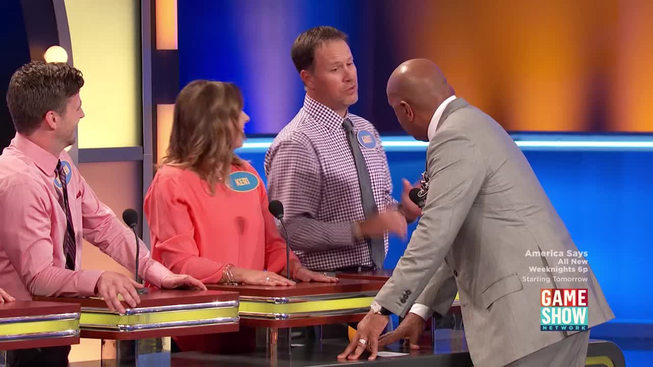 The host leans in, gesturing with his hands as he speaks to a contestant. Across from him, two other contestants look on, one smiling slightly.