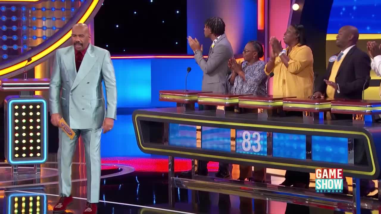 Steve Harvey stands on stage, holding a yellow card, as a family claps behind him. The scoreboard displays "83" in bright blue numbers.