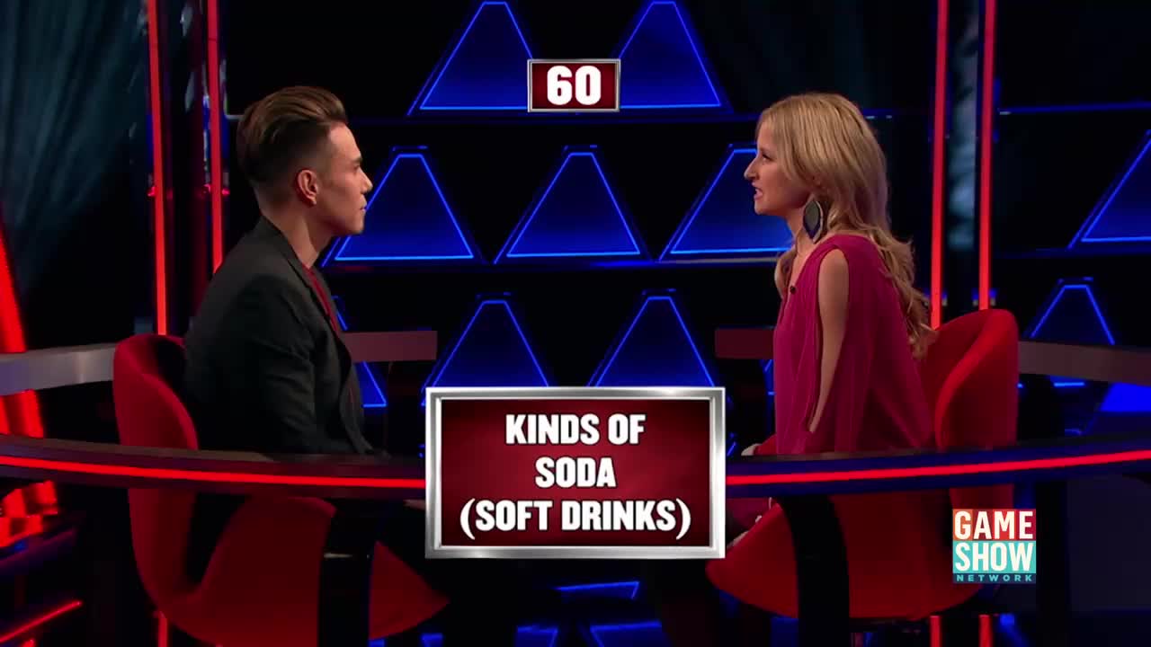 A man and a woman sit across from each other on a game show set. A sign between them reads "KINDS OF SODA (SOFT DRINKS)."