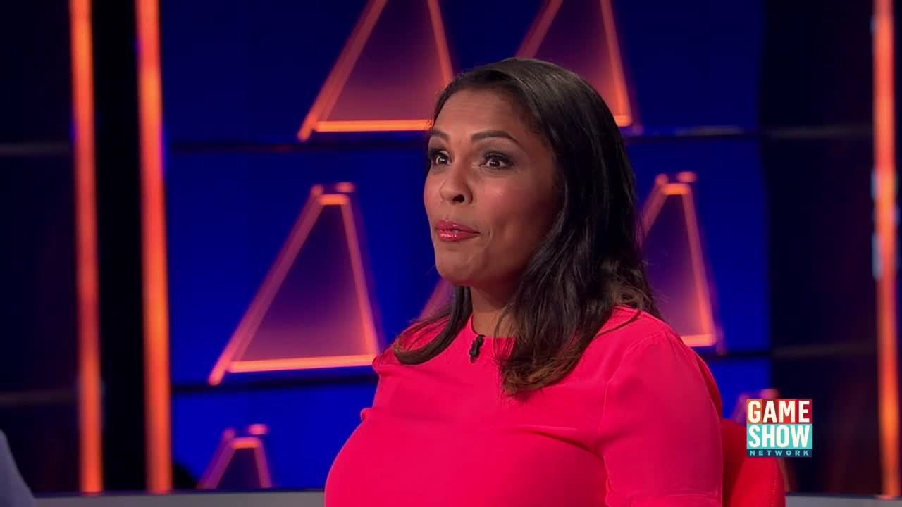 A woman in a bright pink top stands on a game show set. Behind her, illuminated triangles glow on a dark blue backdrop, with the Game Show Network logo visible on the right.