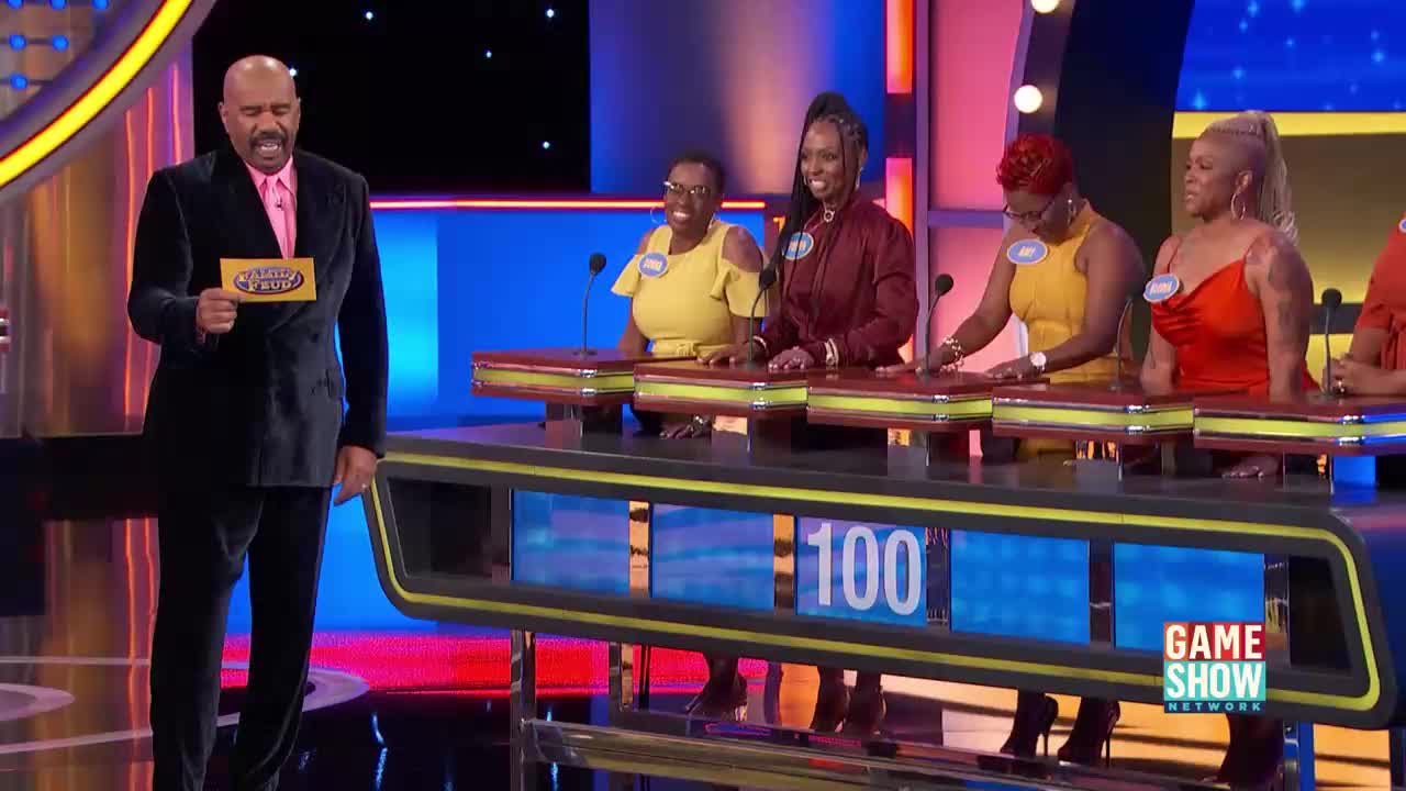 Steve Harvey stands holding a card, facing a line of contestants. The women are gathered at their podiums, ready for the next question.