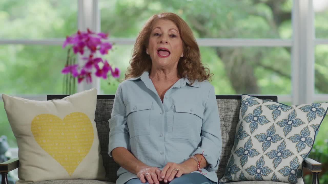 A woman with auburn hair sits on a couch, her mouth open as if speaking. Behind her, a vase of bright pink orchids stands on a table.