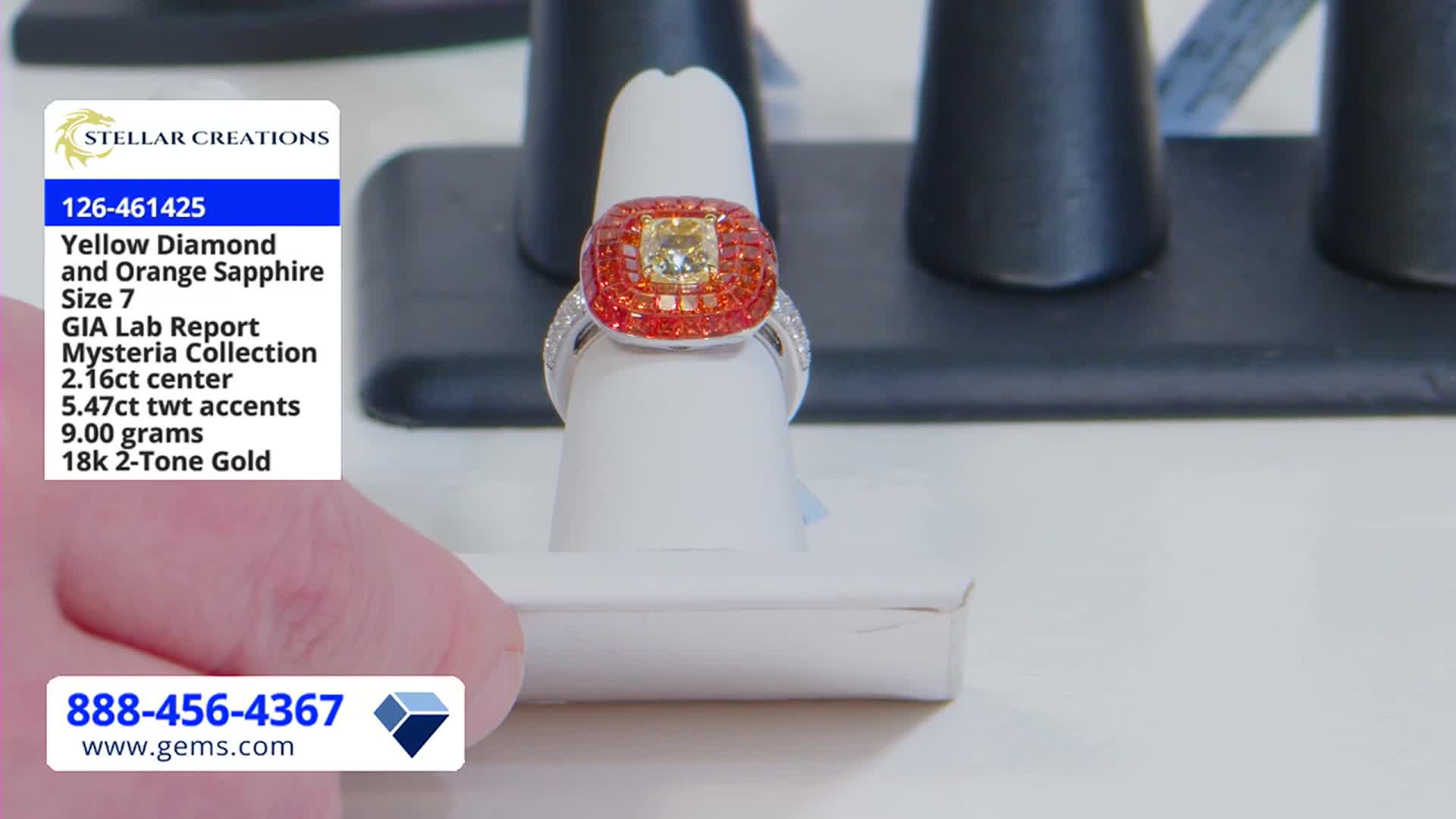 A ring with a yellow diamond center and orange sapphire accents sits on a white display stand. The Gem Shopping Network's phone number and website are visible in the lower corner.
