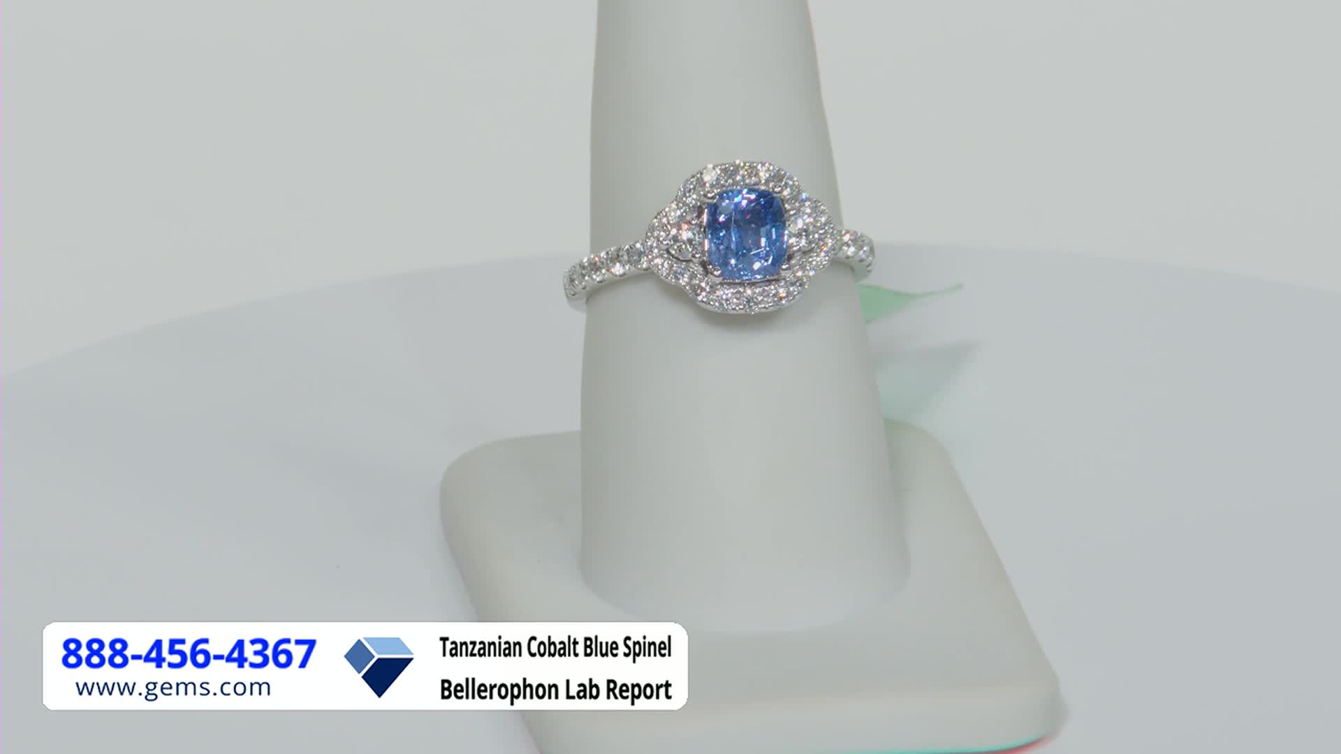A ring with a blue gemstone is displayed on a white stand, likely for a Gem Shopping Network presentation. The ring's band is studded with small diamonds, and the central stone is surrounded by a halo of smaller ones.
