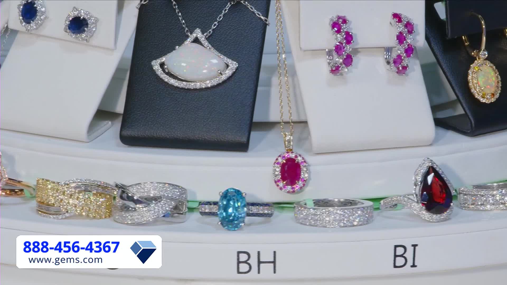 Jewelry glitters under bright studio lights, displayed on white and black pedestals. The Gem Shopping Network logo is visible, along with a phone number for viewers in the United States.
