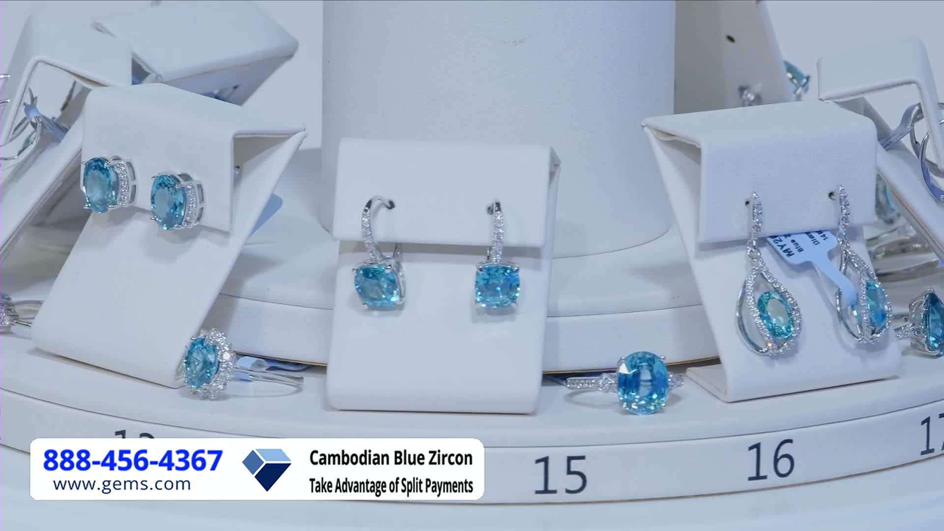Jewelry, including earrings and rings, is displayed on white pedestals, each piece showcasing a vibrant blue stone. The Gem Shopping Network logo and a phone number are visible in the lower left corner.
