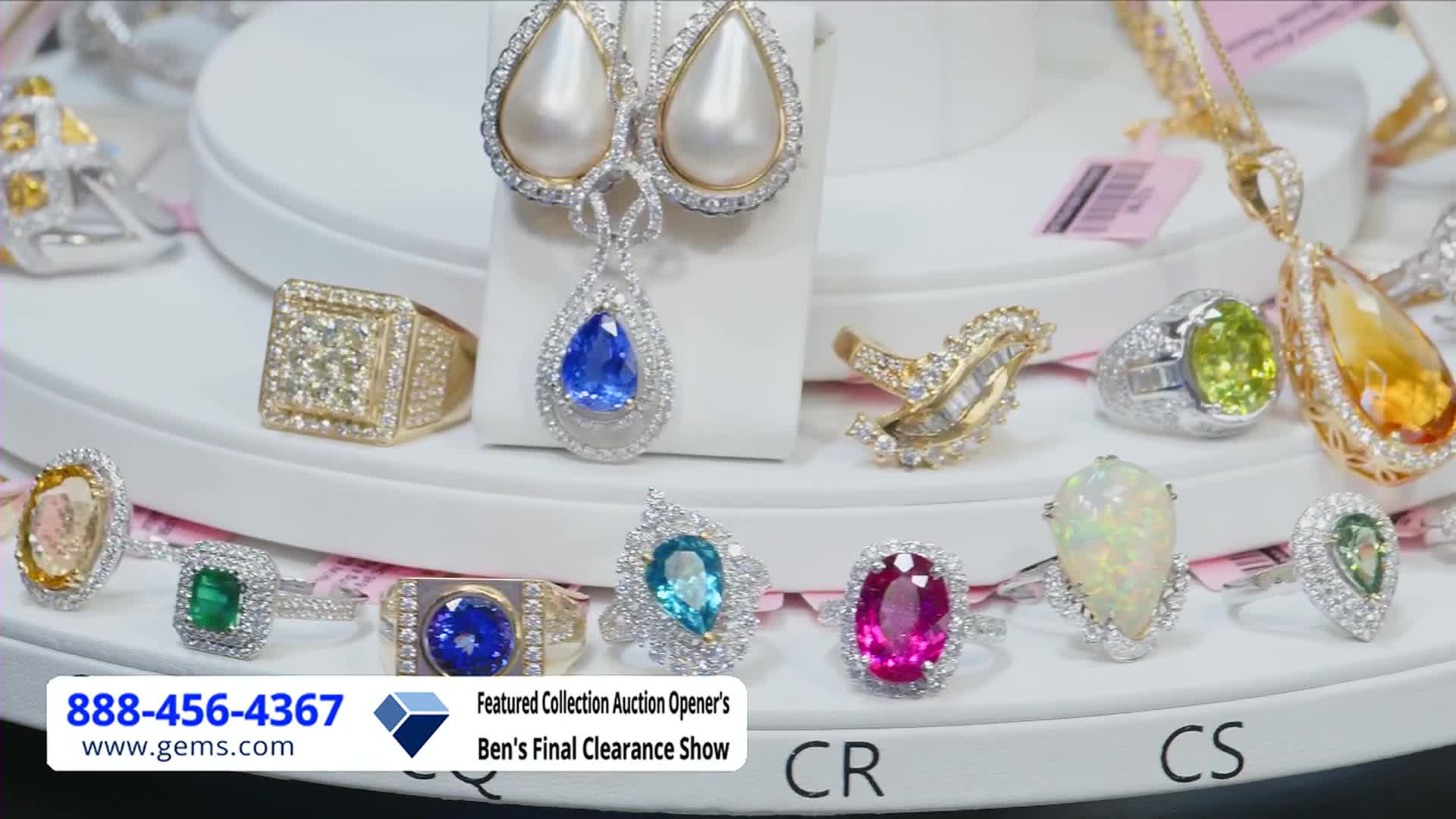 A dazzling array of jewelry is on display, including rings with emeralds, opals, and sapphires, all sparkling under the bright lights. The Gem Shopping Network logo and phone number are visible at the bottom, advertising Ben's Final Clearance Show.
