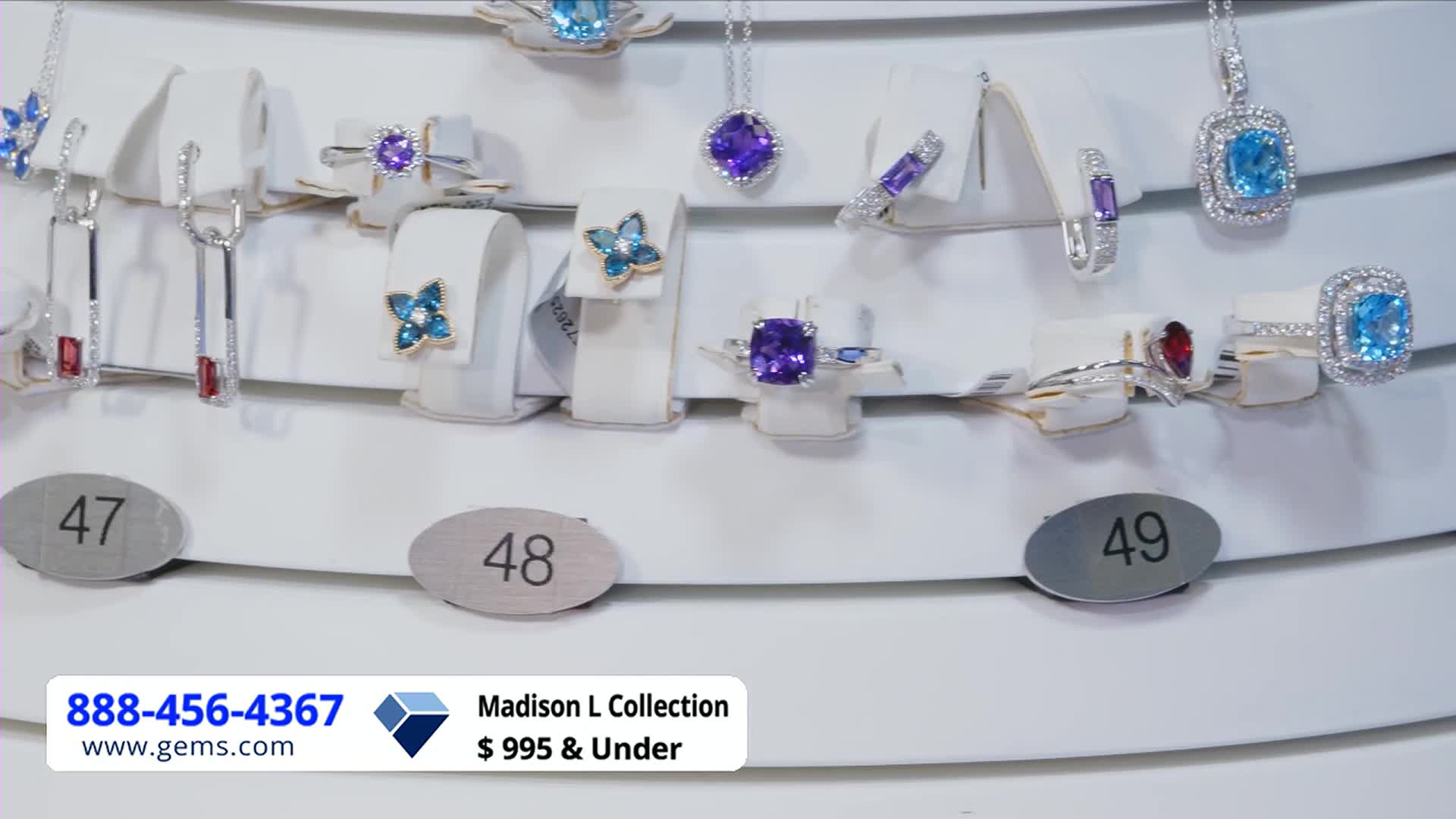 Jewelry, mostly rings and necklaces, are displayed on white stands, each item illuminated by the studio lights. The Gem Shopping Network is running a Madison L Collection sale, with prices under $995.
