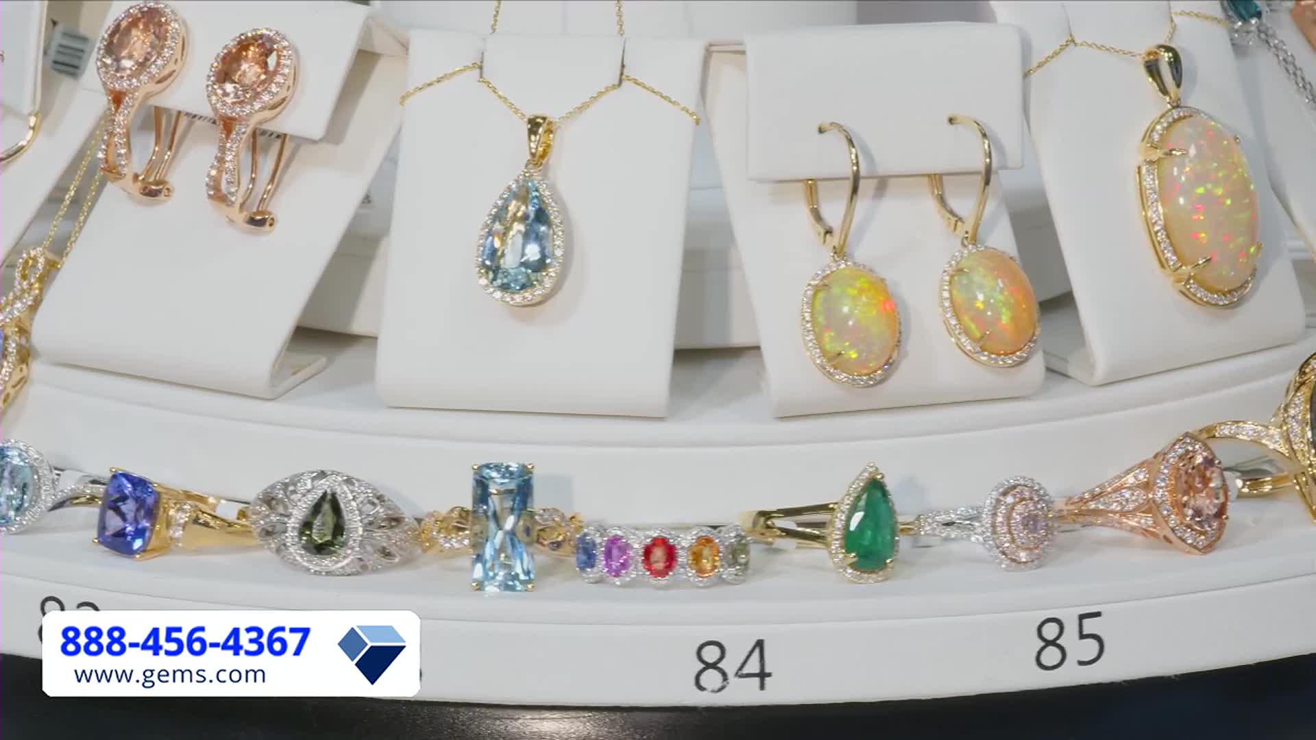 Jewelry, displayed on white stands, gleams under bright lights. A phone number and website address flash in the lower left corner, probably from the Gem Shopping Network.
