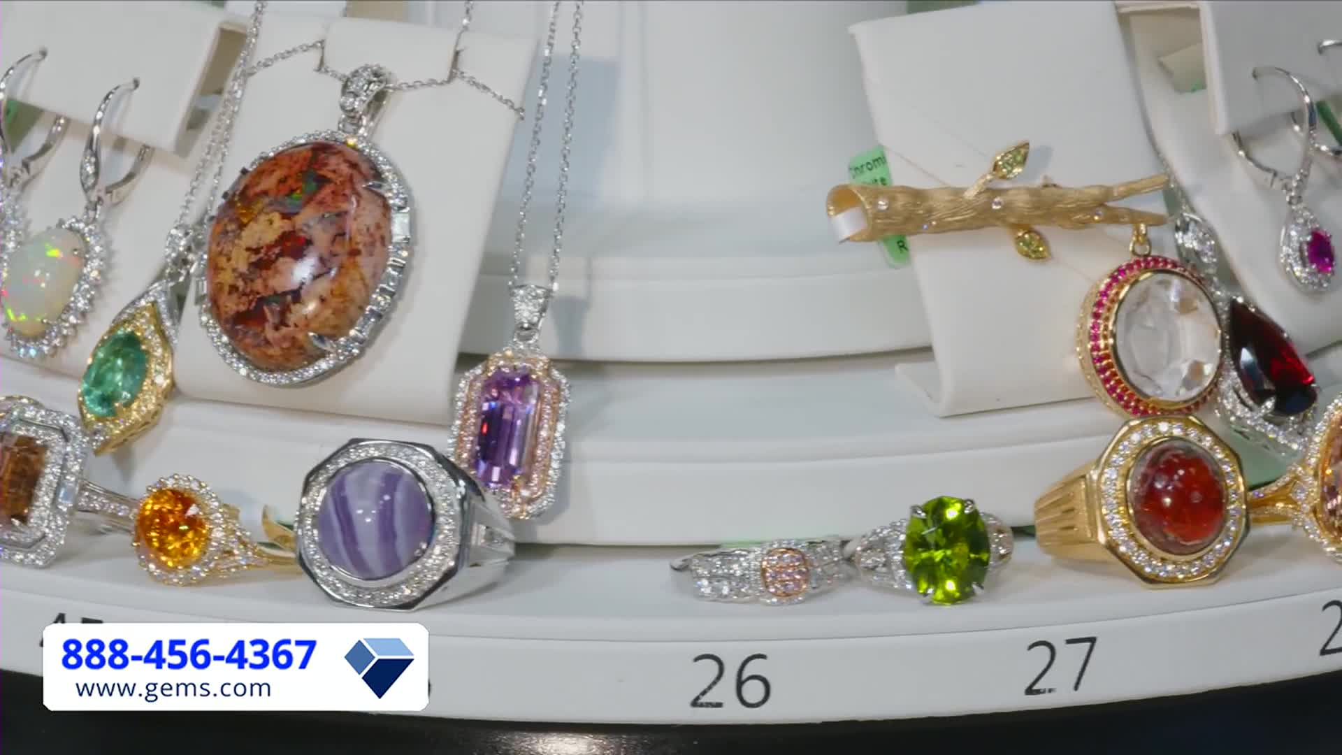 A display of jewelry, including rings and pendants, sits on a white, tiered shelf. The Gem Shopping Network's phone number and website are visible in the lower left corner.
