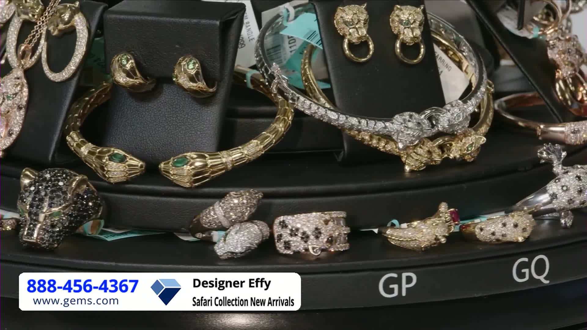 A display of jewelry sparkles under bright lights, likely for a Gem Shopping Network broadcast. Gold snake bracelets and diamond-encrusted rings are arranged on a rotating platform.
