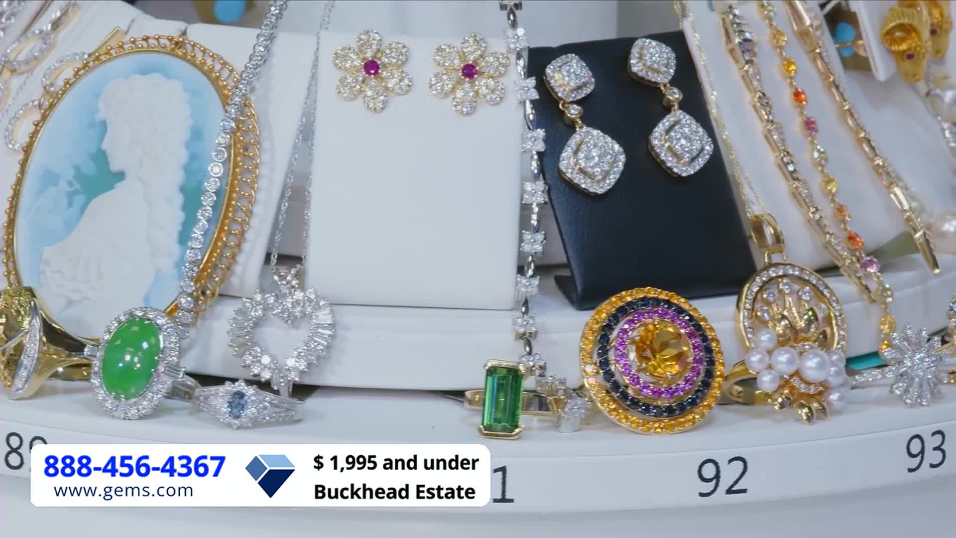 On a rotating display, various pieces of jewelry sparkle under bright lights. A ring with a large, yellow stone surrounded by purple and black gems catches the eye.
