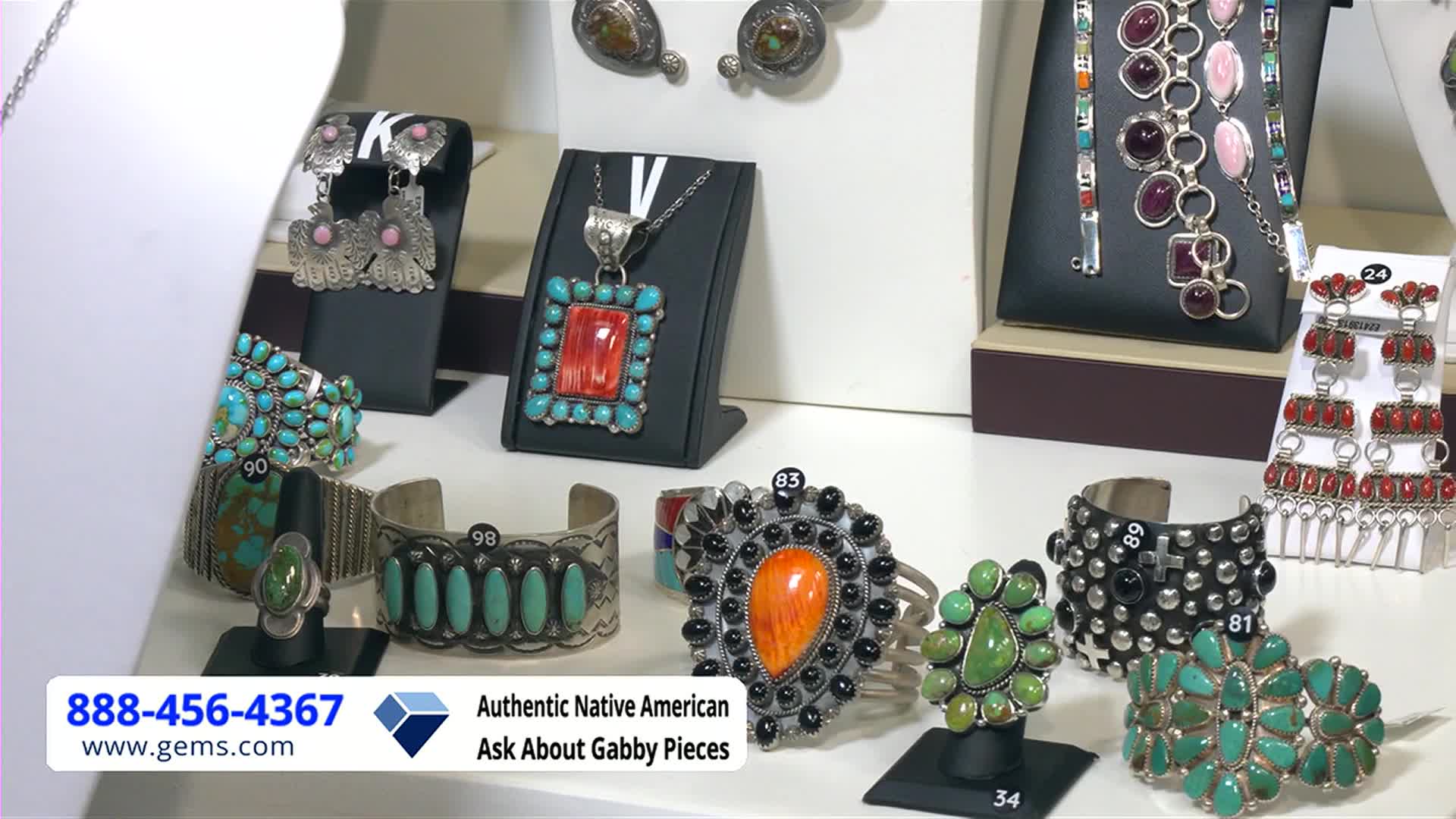 A display case holds a variety of Native American jewelry, from turquoise bracelets to earrings with coral accents. The Gem Shopping Network logo and phone number are in the lower left corner.
